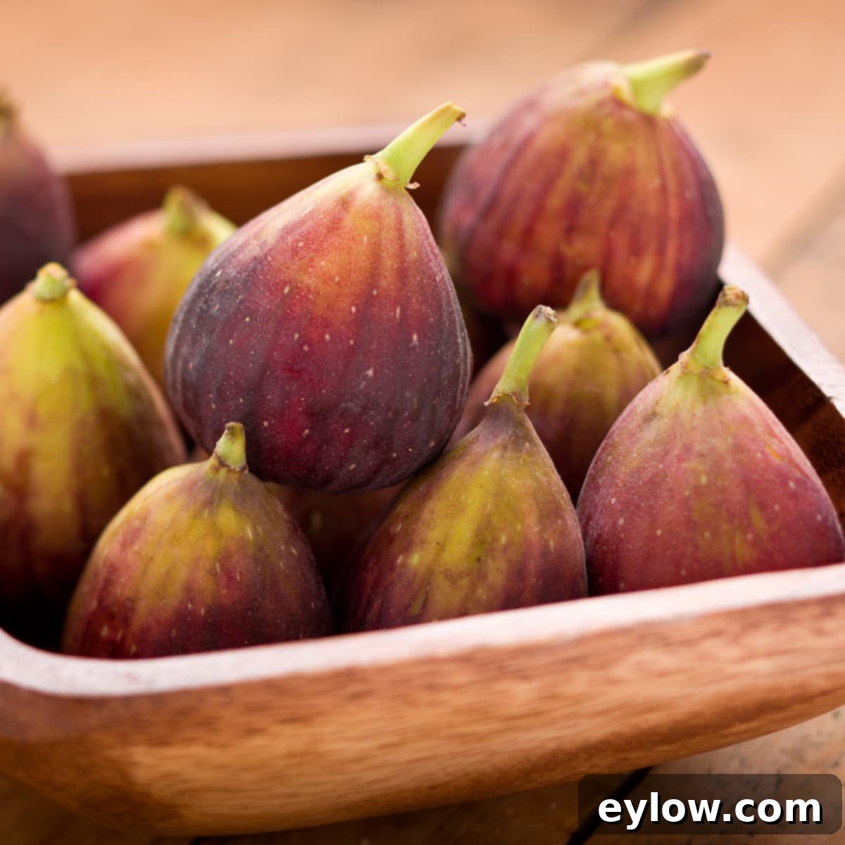 A brown wooden bowl of black mission figs with purplish skin and green tops, showcasing their ripeness.