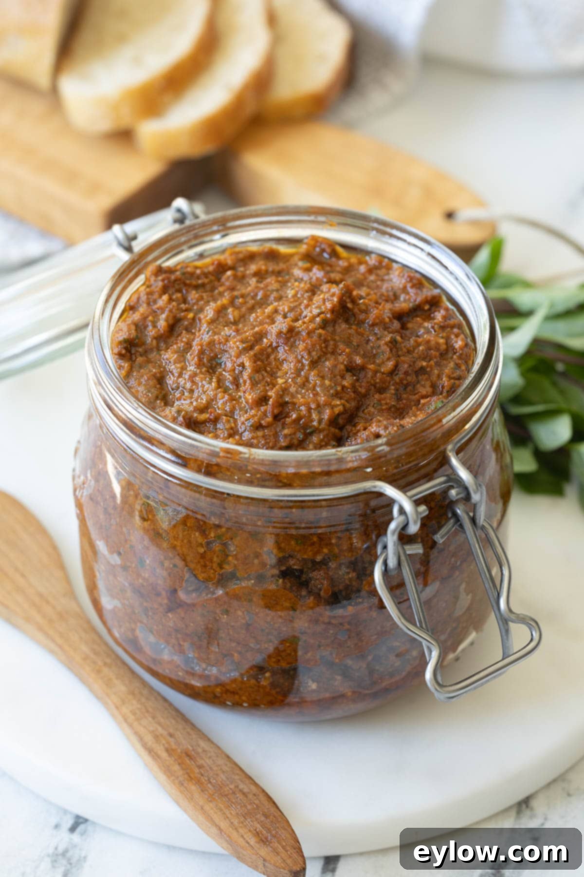 Crimson Sun Pesto 2 An open glass jar of sun-dried tomato pesto with bread slices behind and basil leaves.