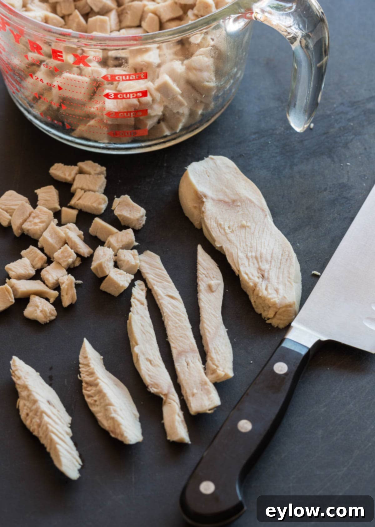 Chopping cooked chicken on a black cutting board into cubes with a chef's knife, ensuring uniform pieces for the salad.