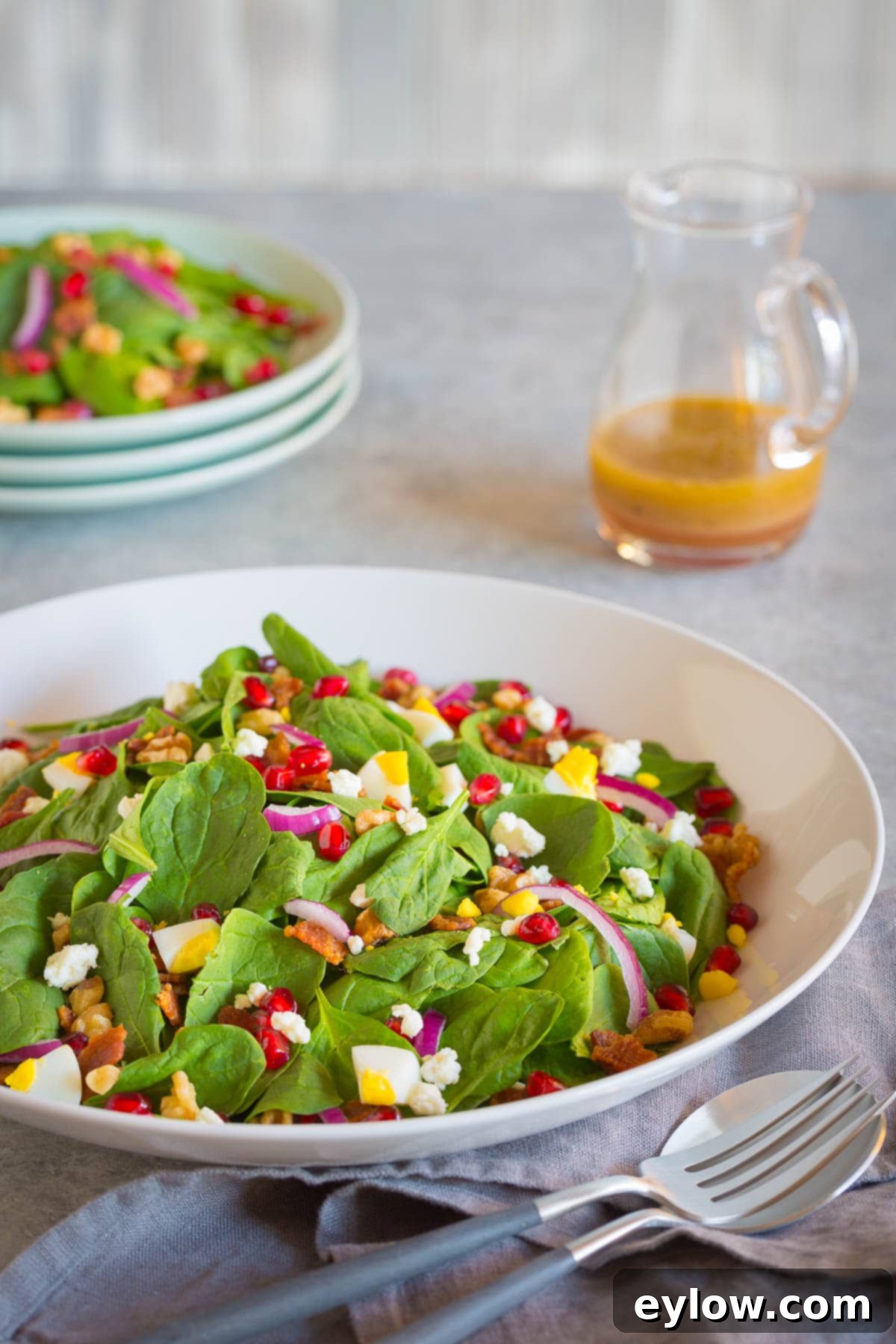 A serving bowl of a baby spinach salad, small plates in the background, and a glass pitcher of dressing.