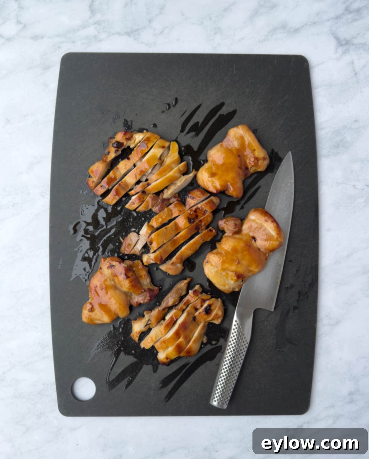 Slicing golden baked chicken thighs on a black cutting board with a chef's knife.