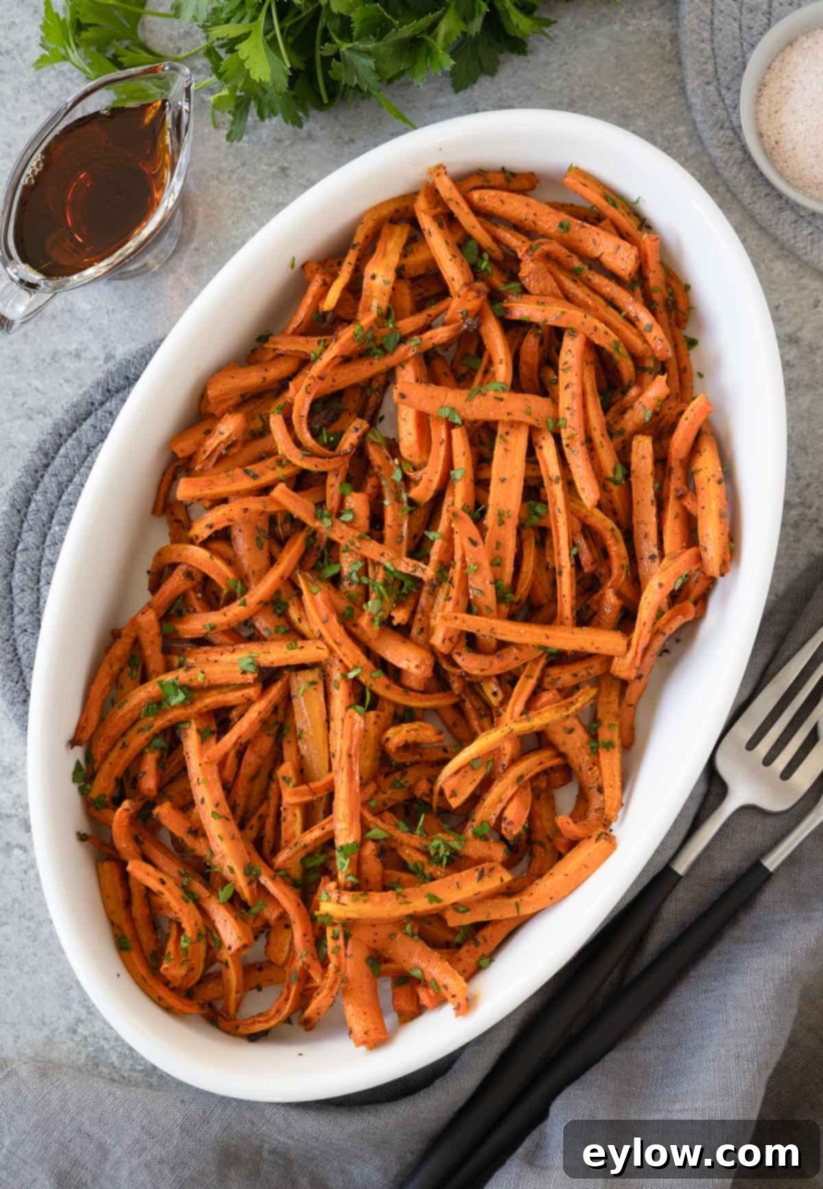 An oval dish of glistening maple roasted carrots with fresh herbs, a small pitcher of maple syrup by the side. 