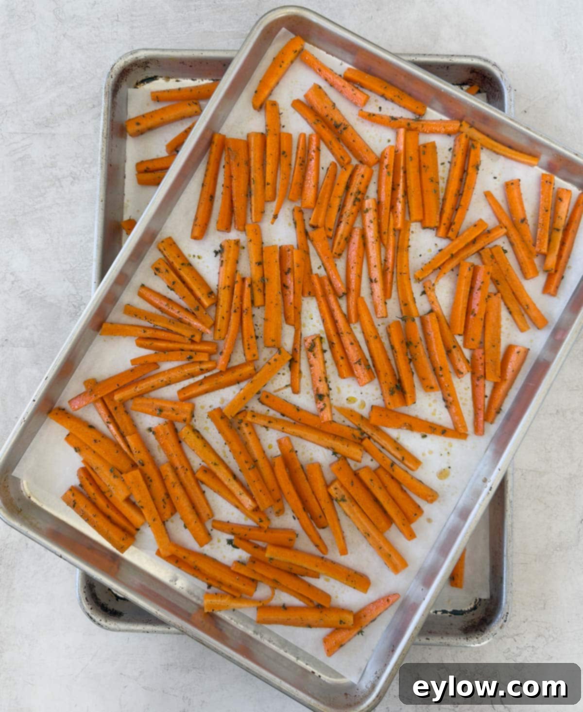 Two trays of seasoned, cut carrots spread in a single layer on baking sheets lined with parchment paper, ready for the oven.