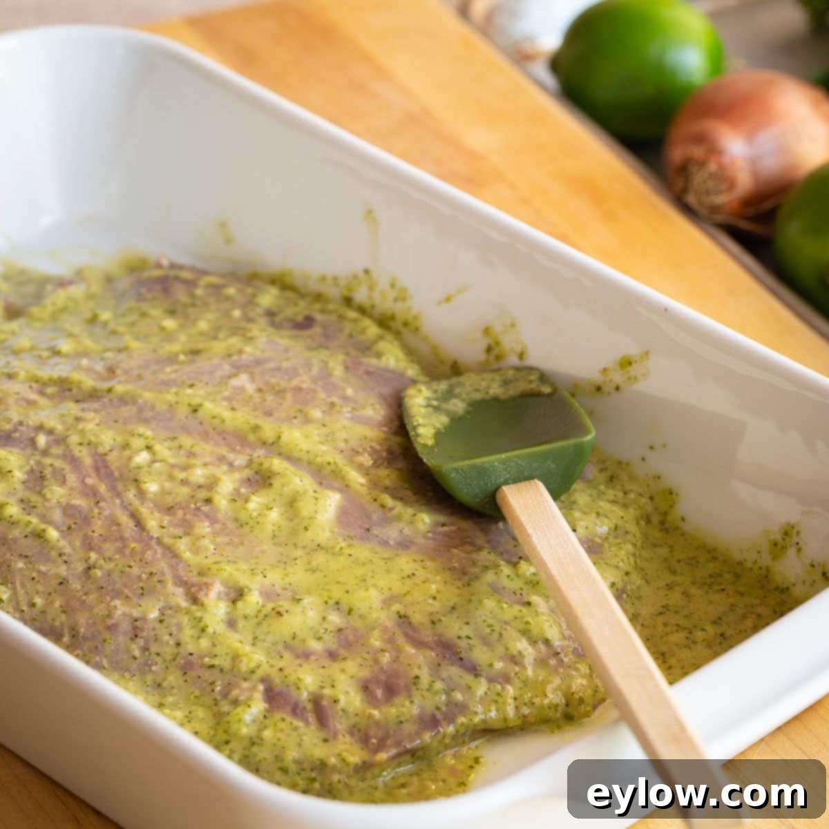 Flank steak marinating in a white casserole dish with a spatula.