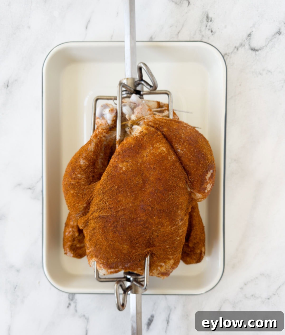 Whole chicken skewered on a rotisserie spit and generously covered with a vibrant reddish-orange seasoning rub, ready for the grill.