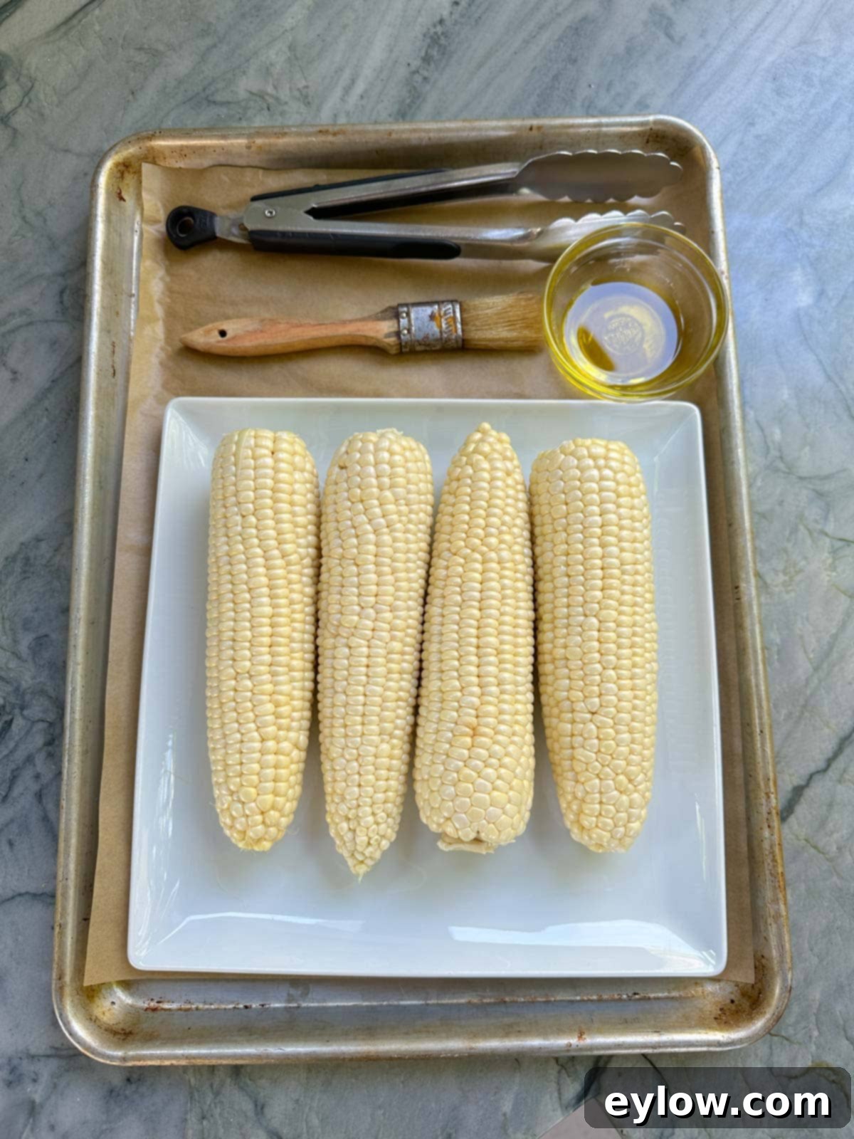 A tray with tongs, pastry brush, olive oil and the shucked corn cobs for grilling.