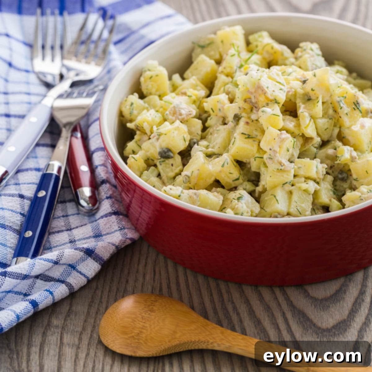Closeup of creamy French potato salad in a serving bowl with a wooden spoon, showcasing fresh herbs and capers.