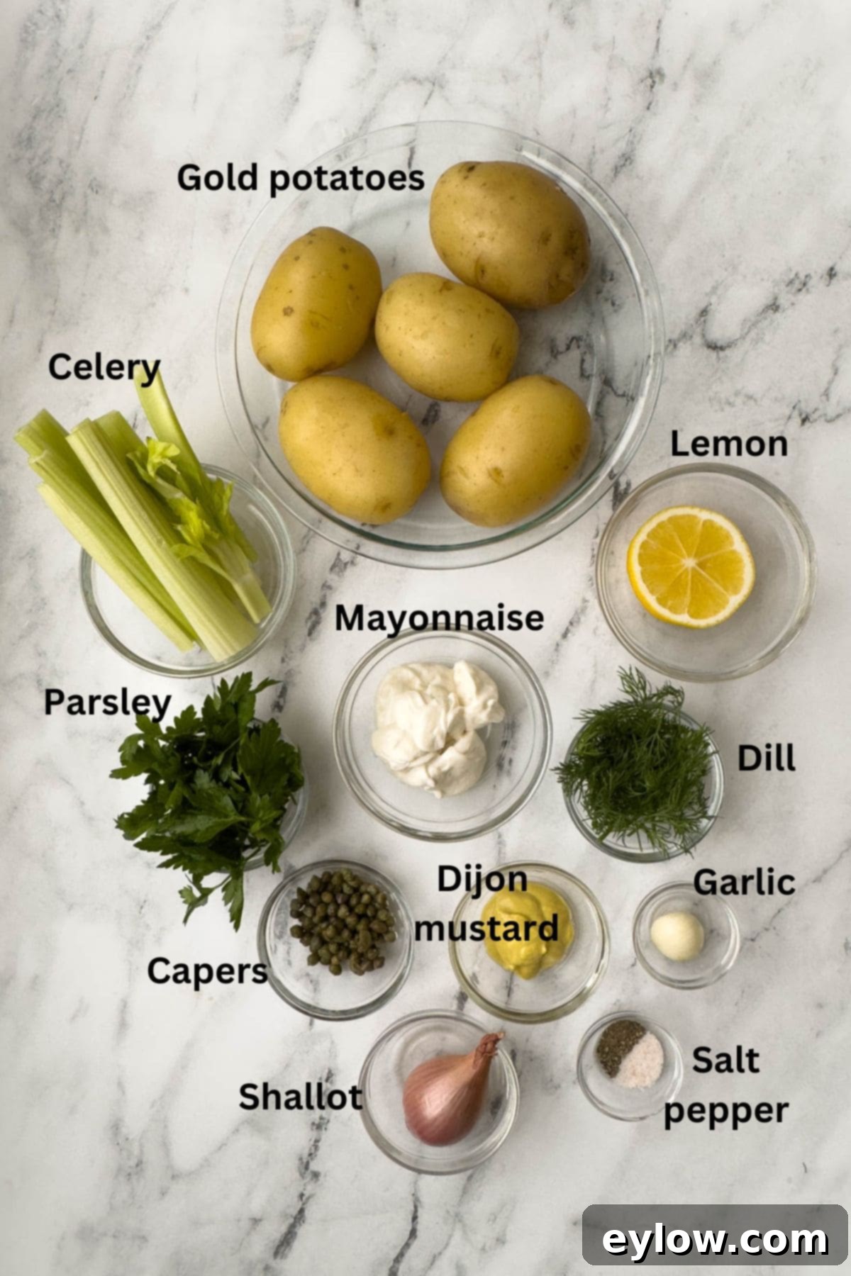 Bowls of fresh ingredients for French potato salad, including chopped shallots, capers, fresh herbs, and Dijon mustard, laid out on a marble counter.