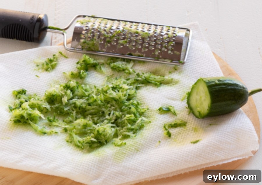 A hand grating a fresh cucumber onto several layers of paper towels using a small silver handheld grater, demonstrating the crucial step of draining excess moisture from the cucumber for creamy tzatziki.