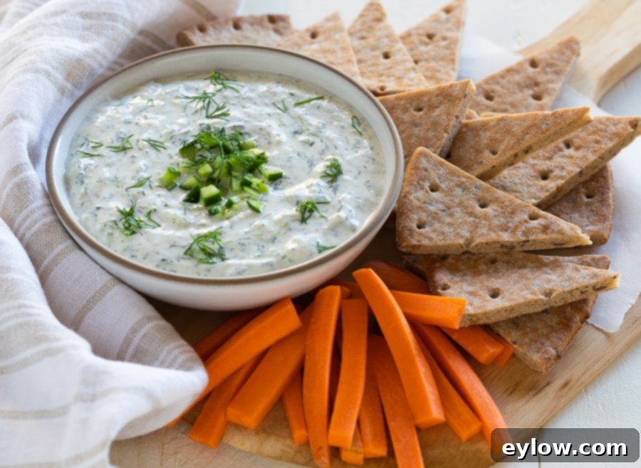 A beautifully presented bowl of homemade Greek tzatziki sauce, garnished with fresh herbs and olive oil, surrounded by various dipping options including cucumber slices, carrot sticks, and warm pita bread on a rustic wooden board. This image highlights its versatility as a dip and appetizer.