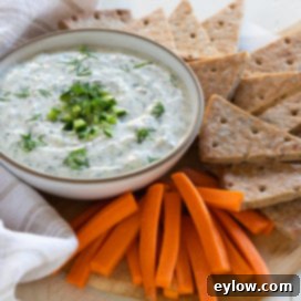 A beautifully presented bowl of homemade Greek tzatziki sauce, garnished with fresh herbs and olive oil, surrounded by various dipping options including cucumber slices, carrot sticks, and warm pita bread on a rustic wooden board. This image highlights its versatility as a dip and appetizer.