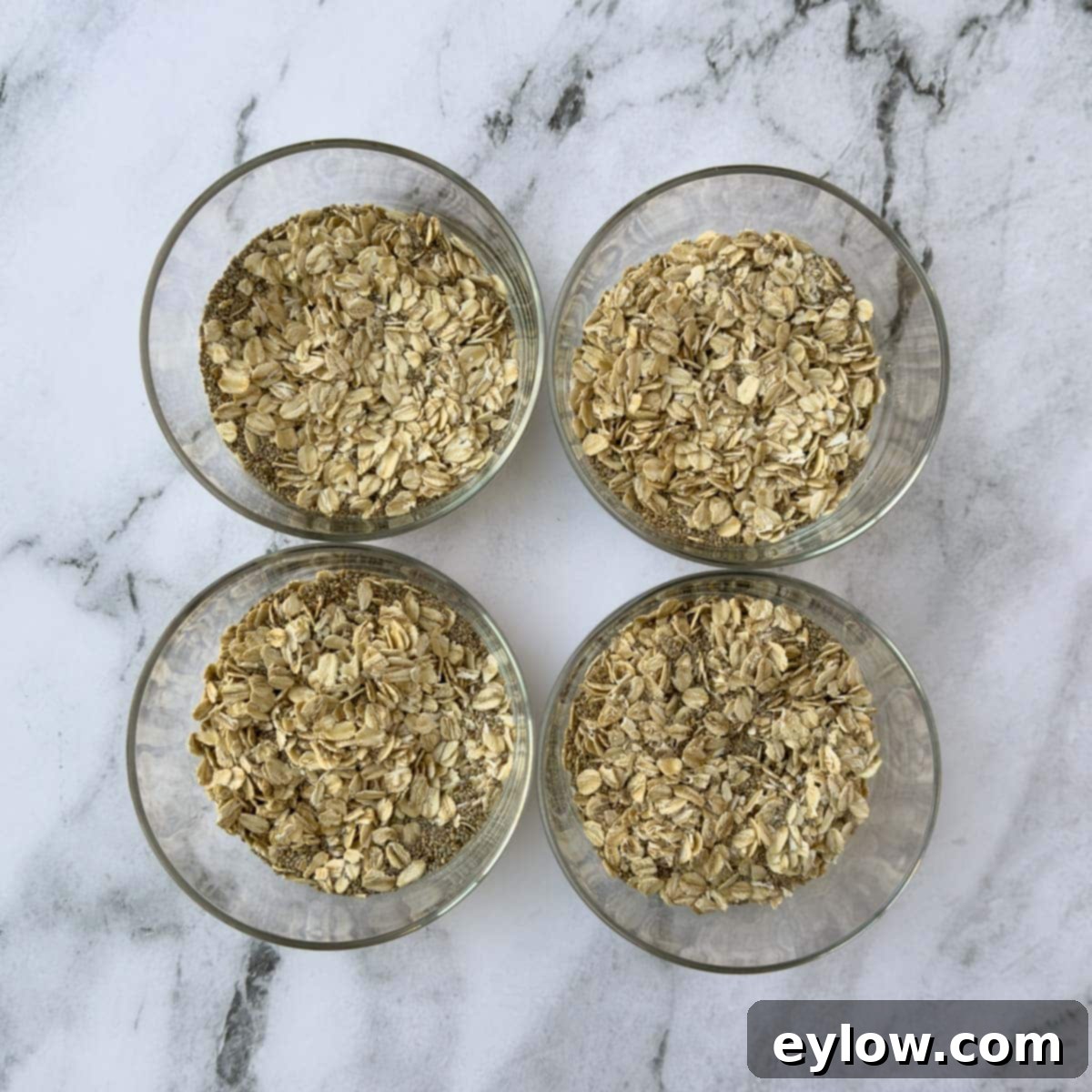 Four glass bowls with dry rolled oats and chia seeds ready for milk and vanilla to be added.