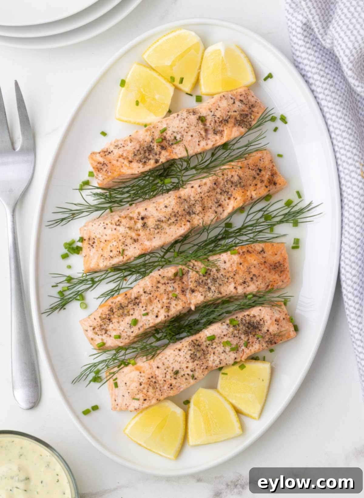 A white oval platter of perfectly cooked, pink steamed salmon fillets garnished with fresh lemon slices and aromatic herbs like dill and parsley.