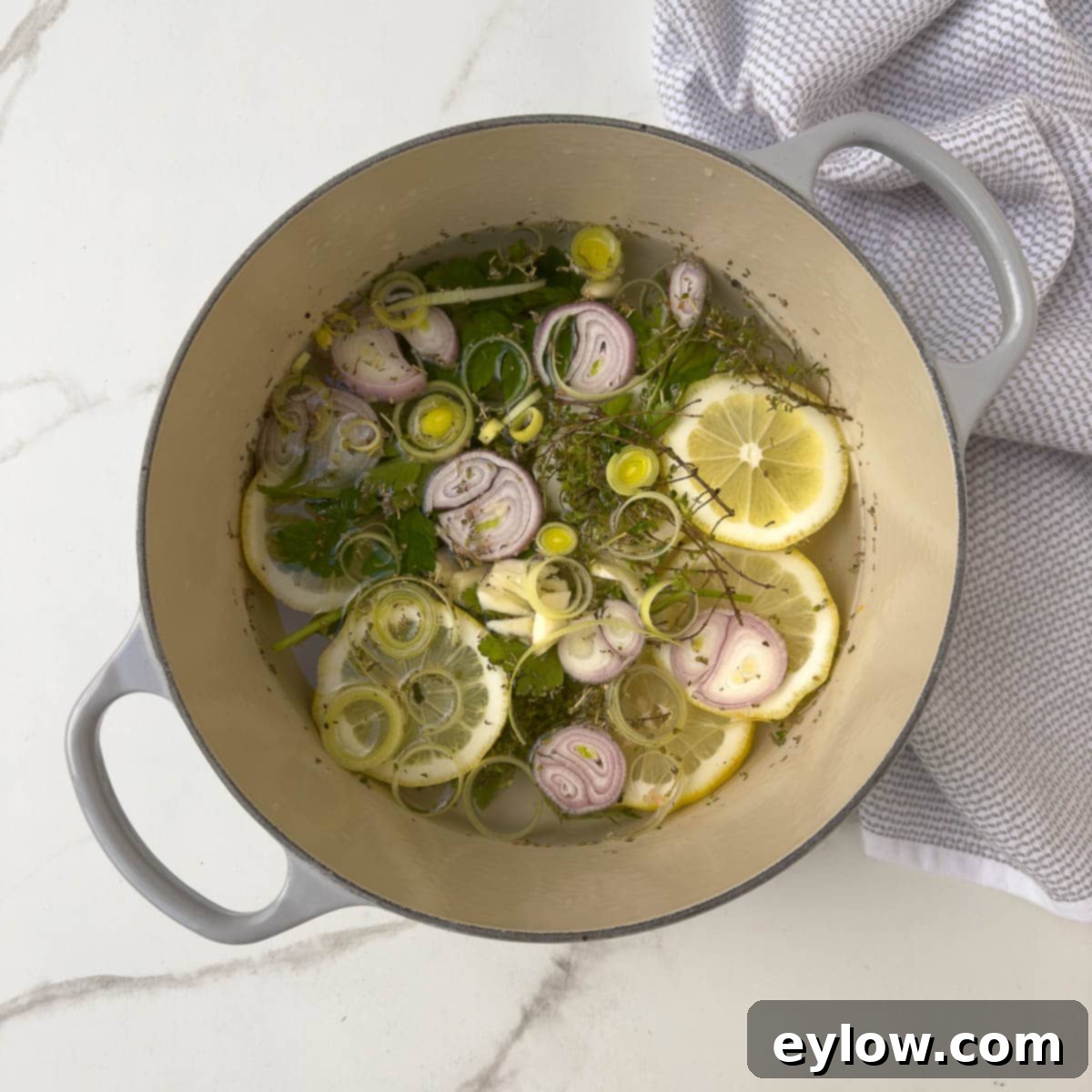 A pot filled with aromatic steaming liquid, featuring slices of fresh lemon, sprigs of green herbs, and chopped shallots, ready to infuse salmon with flavor.
