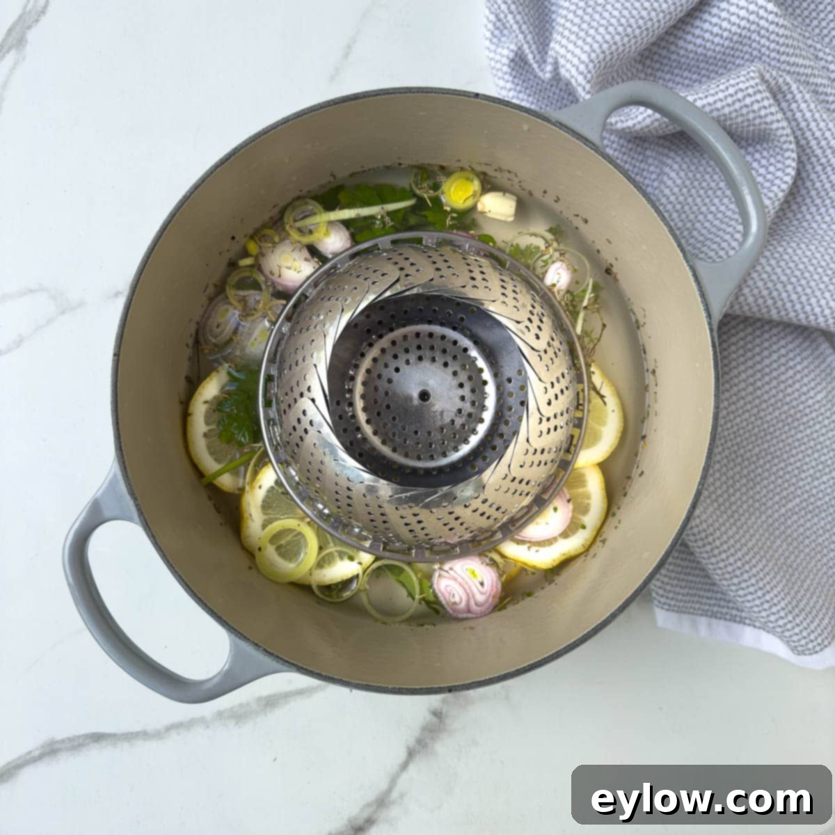 A stainless steel folding steaming rack placed inside a pot containing aromatic liquid for steaming fish, with lemon and herbs visible.