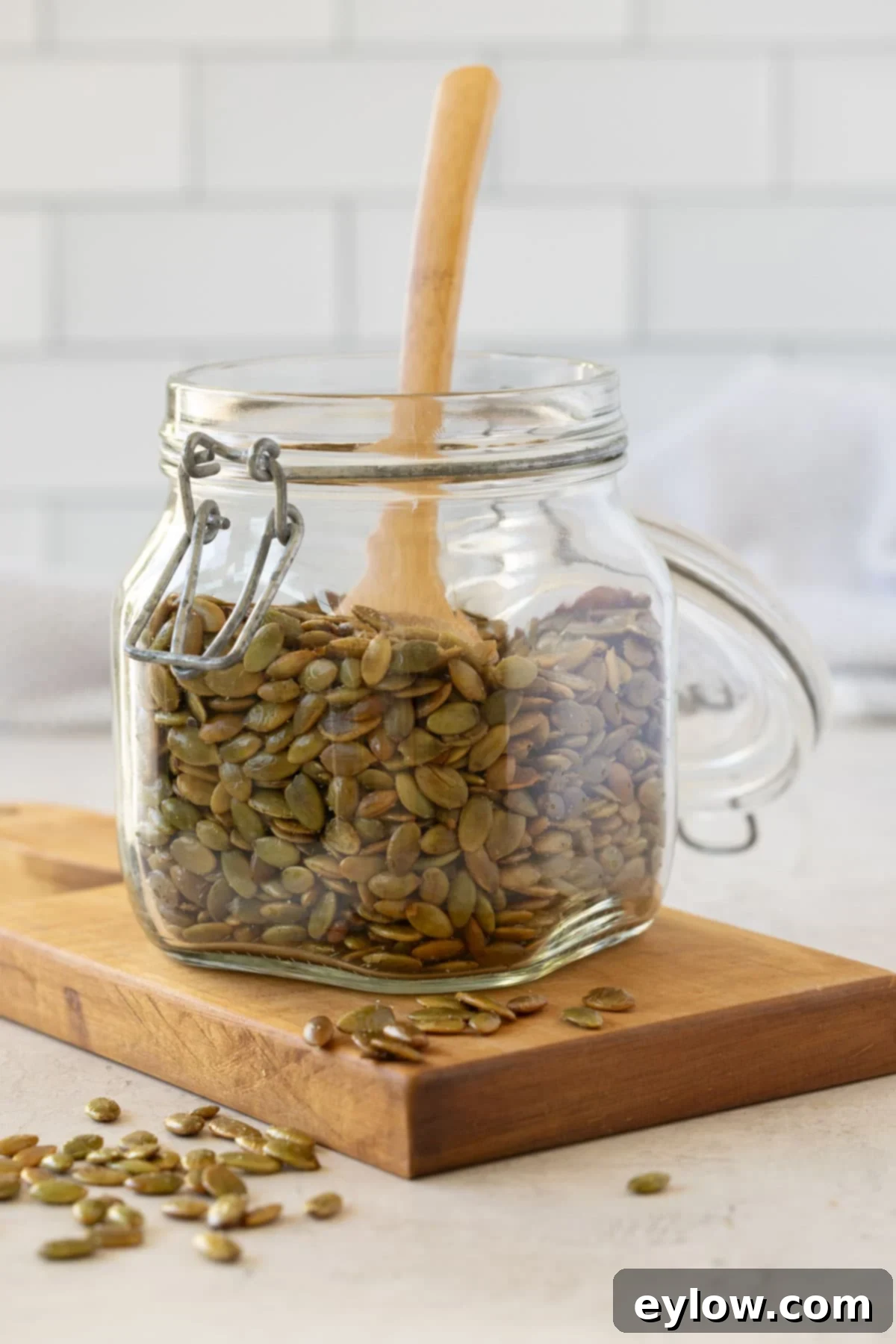 A glass jar on a mini cutting board full of dark green roasted pepitas pumpkin seeds.