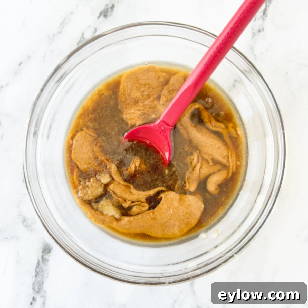 Almond butter and other ingredients for Asian dipping sauce in a glass bowl before blending with a red spatula. All components are distinct.