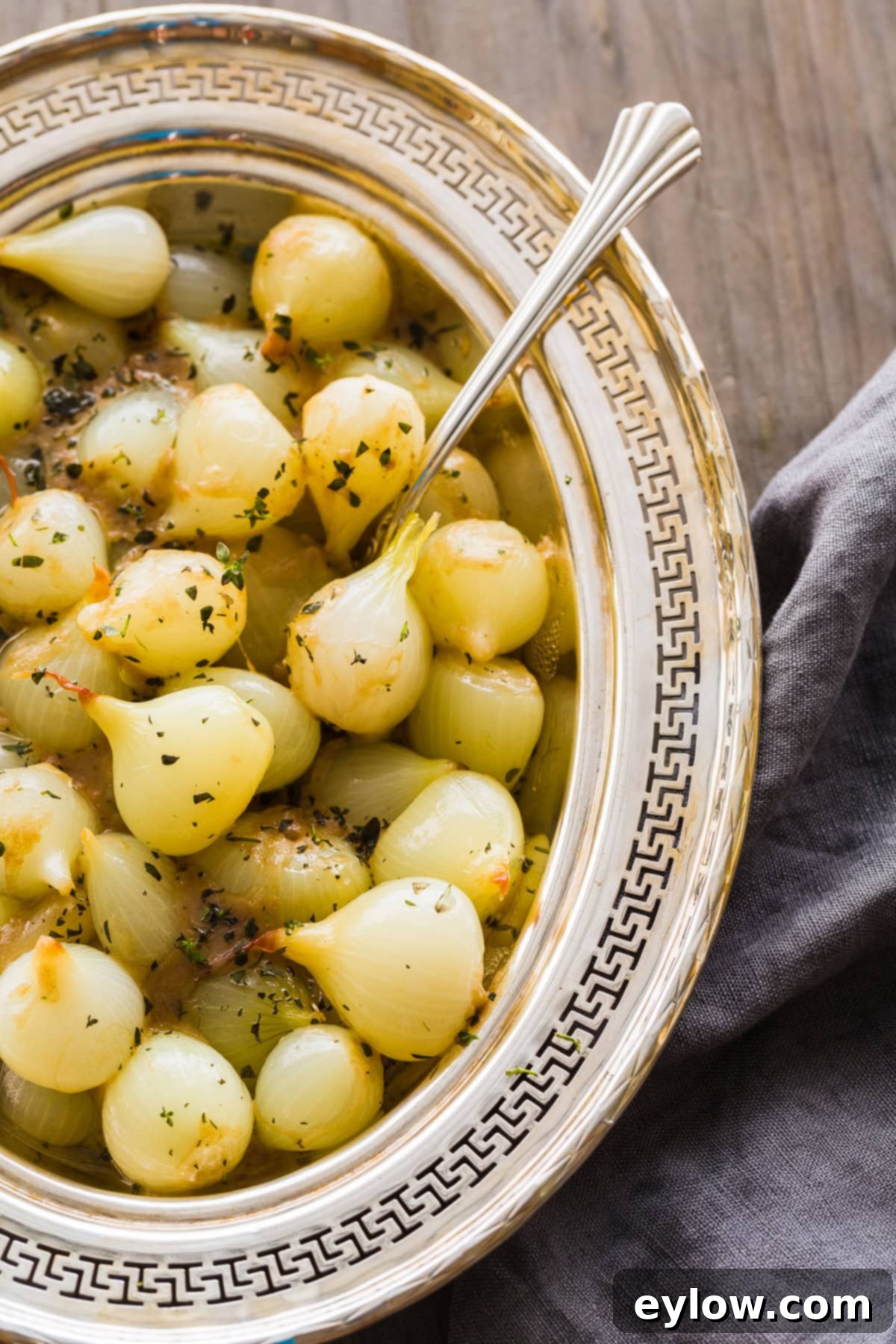 Effortless and Velvety Creamed Pearl Onions 2 An oval silver serving dish filled with perfectly creamed pearl onions, with a silver spoon resting in the dish, ready to be served, garnished with fresh herbs.
