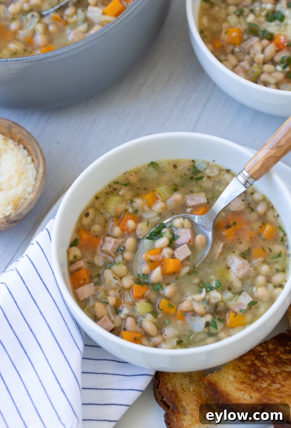 A steaming bowl of navy bean soup with ham and mixed vegetables, garnished with fresh herbs, served on a rustic wooden surface.