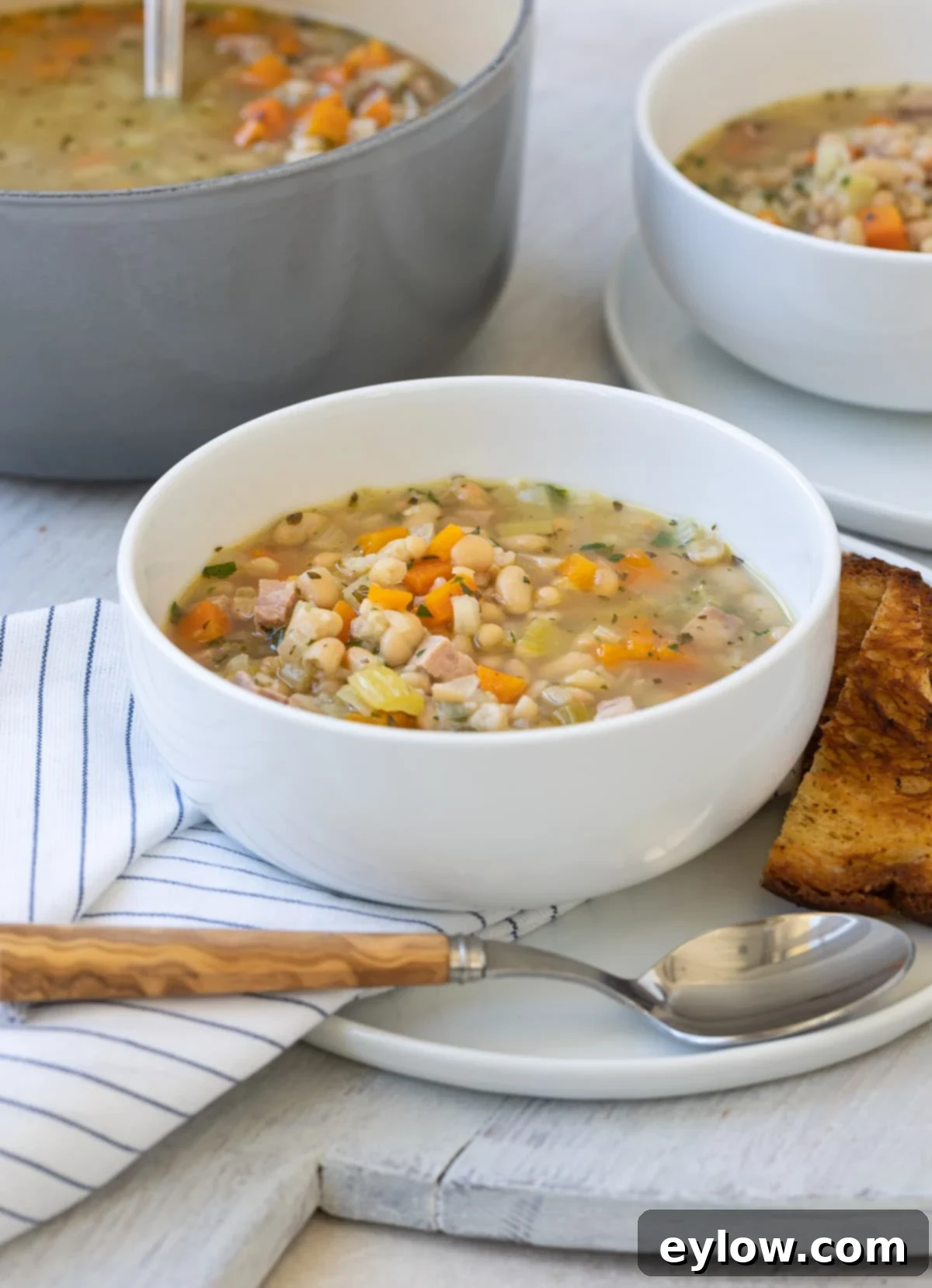 Two bowls of navy bean soup, garnished with parsley, on a table with fresh ingredients.