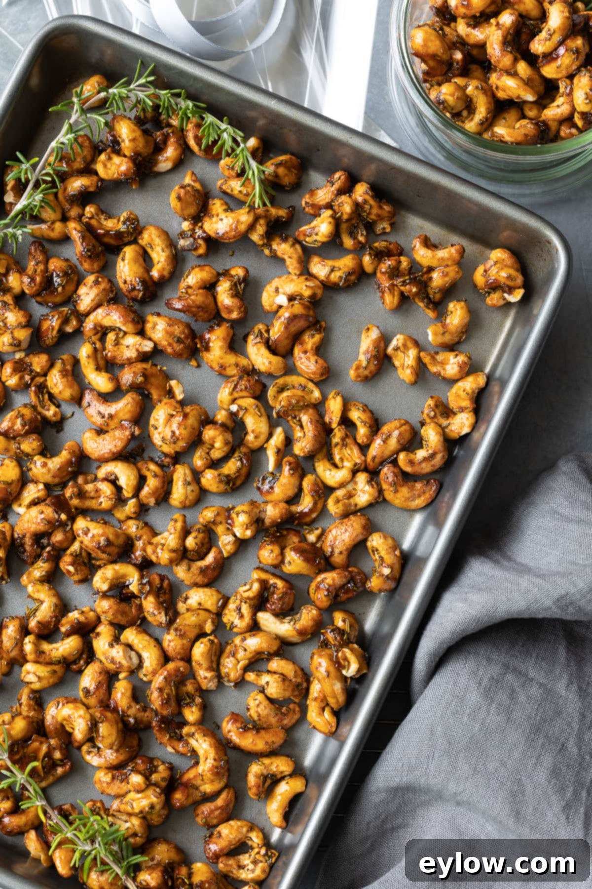 Maple Glazed Sweet and Spicy Cashews 2 Maple roasted cashews on baking sheet with rosemary. A close-up shot shows the golden-brown cashews glistening with maple glaze and flecked with fresh green rosemary leaves.