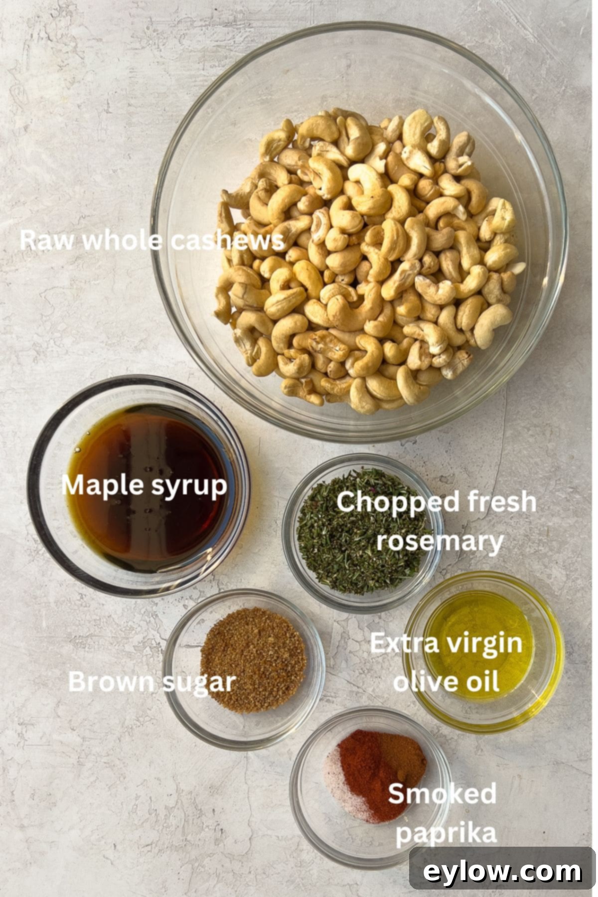 Maple Glazed Sweet and Spicy Cashews 3 Roasted cashews ingredients, labeled, on counter. The image displays raw cashews, a bottle of maple syrup, a sprig of fresh rosemary, olive oil, brown sugar, vanilla extract, paprika, chipotle powder, and sea salt.