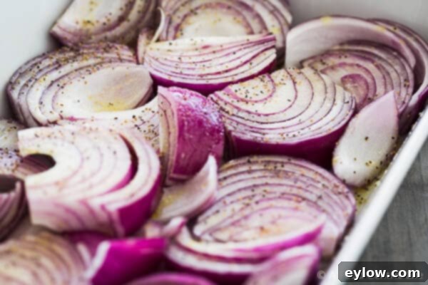 Close-up view of vibrant red onion wedges in a large baking dish, tossed with olive oil and spices, ready for roasting.