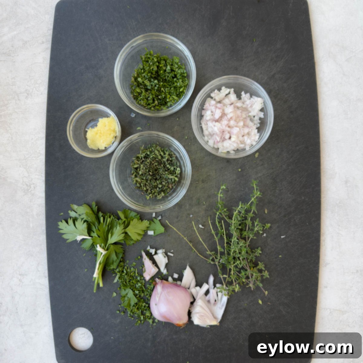 A black cutting board with chopped herbs, shallots and garlic.
