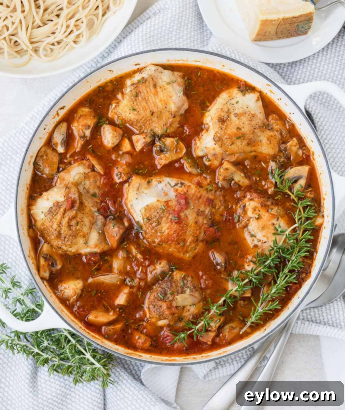Effortless One Pan Italian Chicken Thighs 2 A white pan full of braised chicken thighs in red sauce with herbs, mushrooms, rosemary sprigs.
