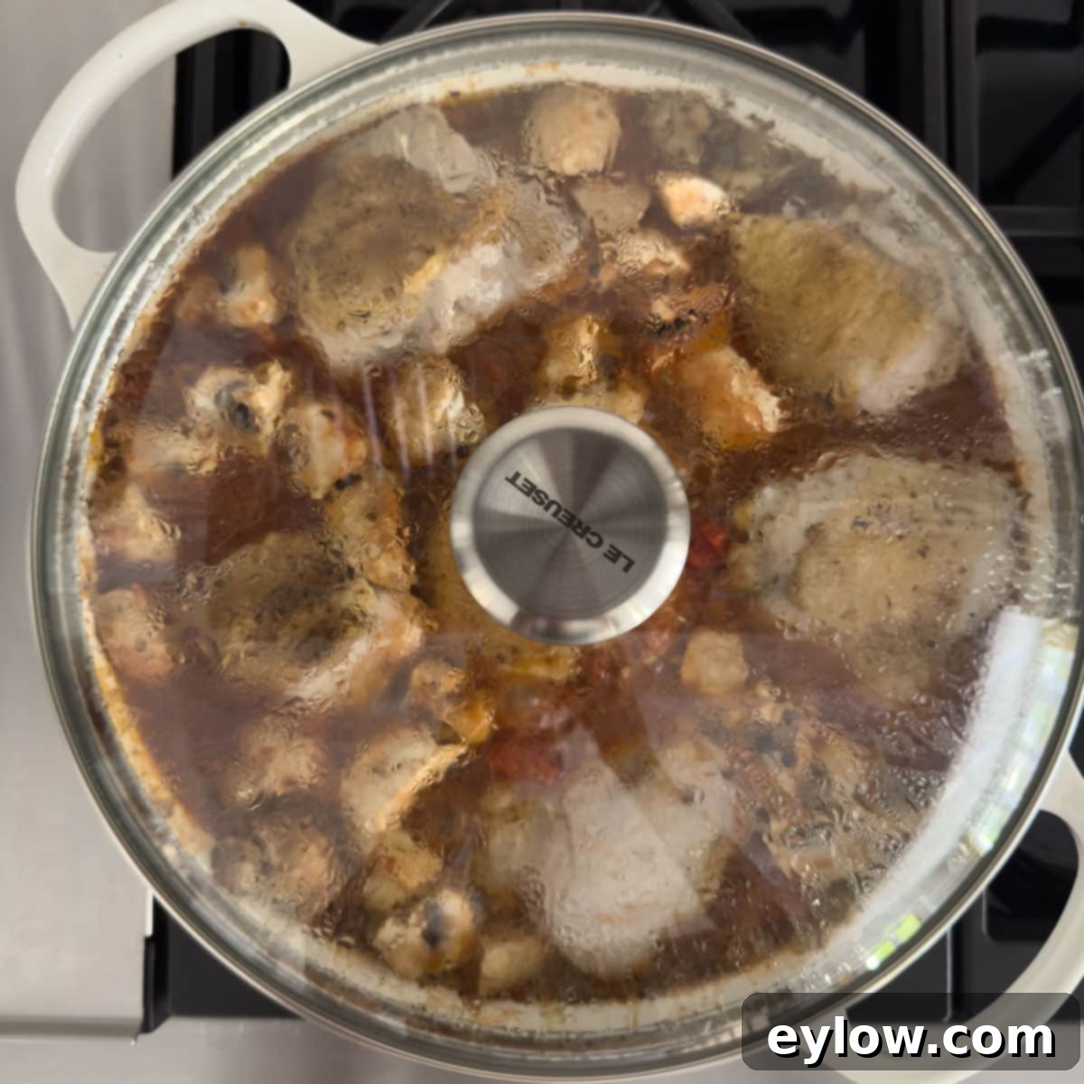 Effortless One Pan Italian Chicken Thighs 10 White pan with a glass lid on the stovetop cooking chicken thighs and vegetables.