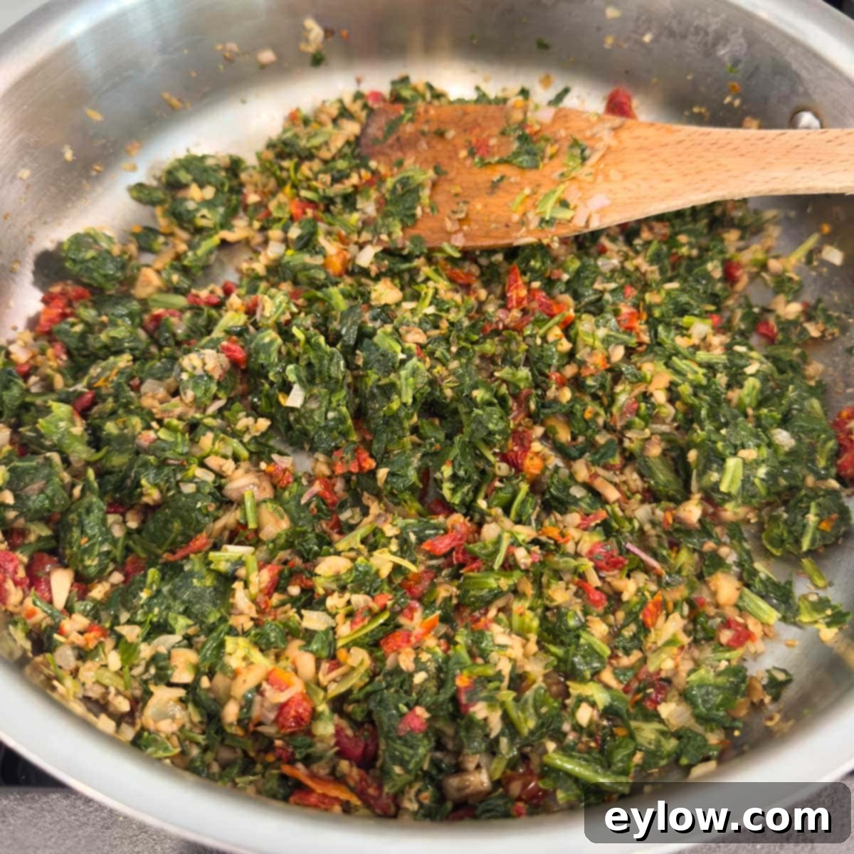 The savory filling for spinach stuffed mushrooms, a vibrant mixture of sun-dried tomatoes, spinach, and Italian seasoning, cooking in a stainless steel sauté pan.
