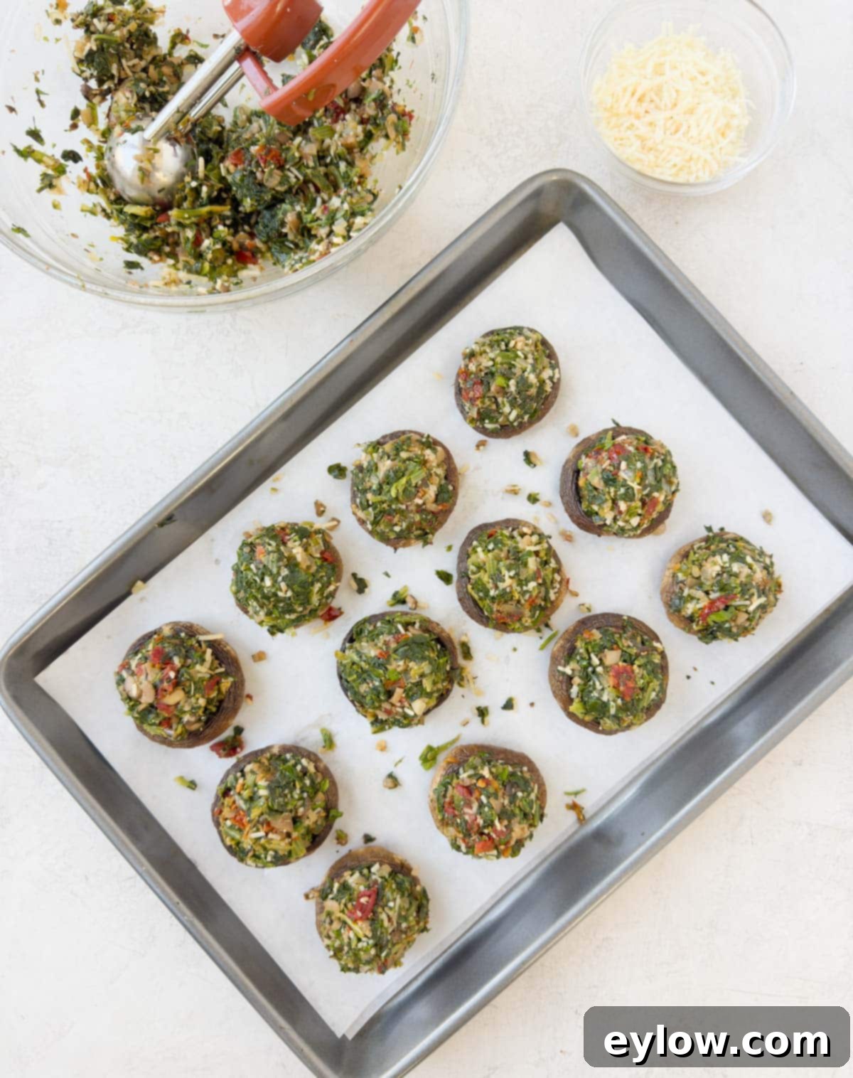 A baking tray with partially stuffed mushrooms, a bowl of filling, and a pile of Parmesan cheese on the side, demonstrating the stuffing process.