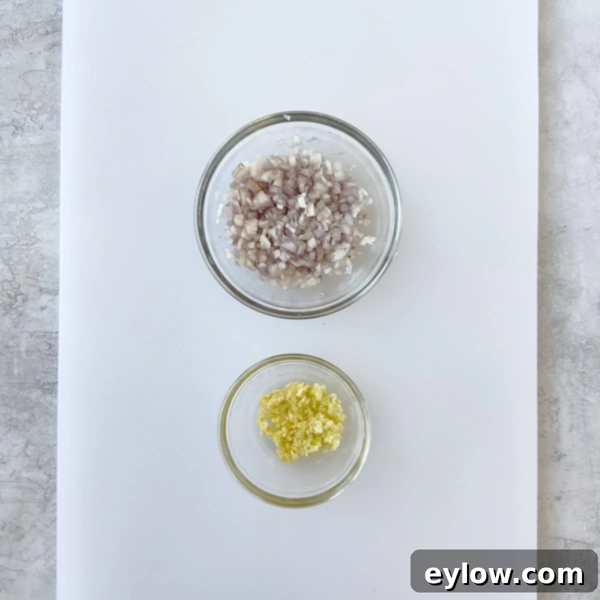 Finely chopped shallot and garlic in small glass prep bowls on a white cutting board, showcasing mise en place.