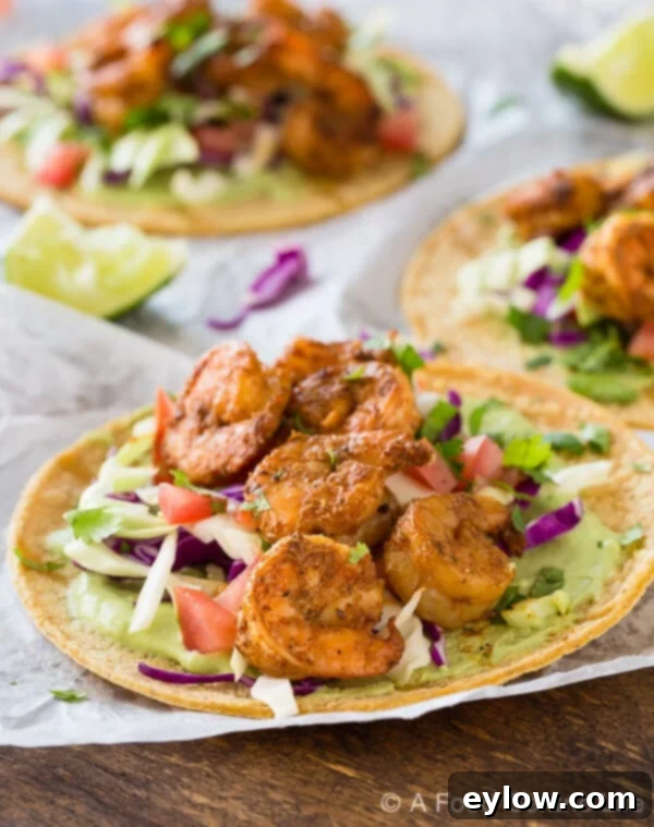 Grilled shrimp tacos piled on white paper on a wood table, with avocado sauce and veggies.