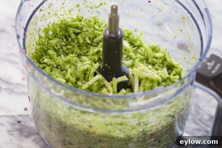 Creamy Apple Cider Crunch Slaw 3 Broccoli florets and stems freshly chopped for broccoli slaw in a food processor bowl, highlighting the ease of preparation.