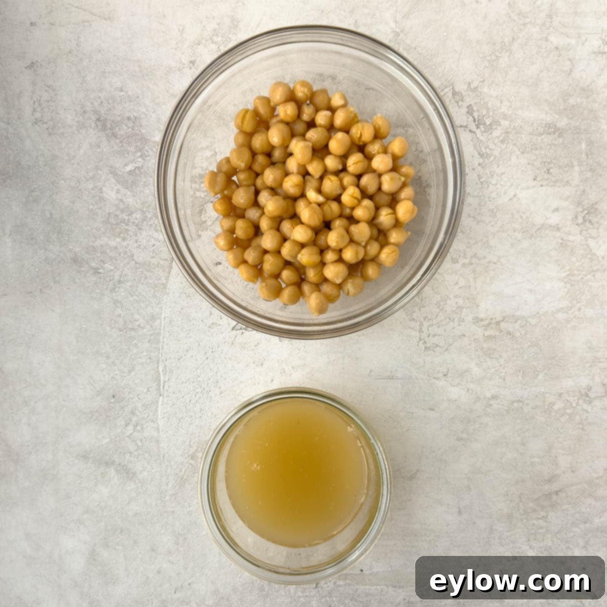 A clear glass bowl holding perfectly drained garbanzo beans, with a separate bowl underneath to collect the valuable aquafaba (canned liquid) for later use in the hummus recipe.