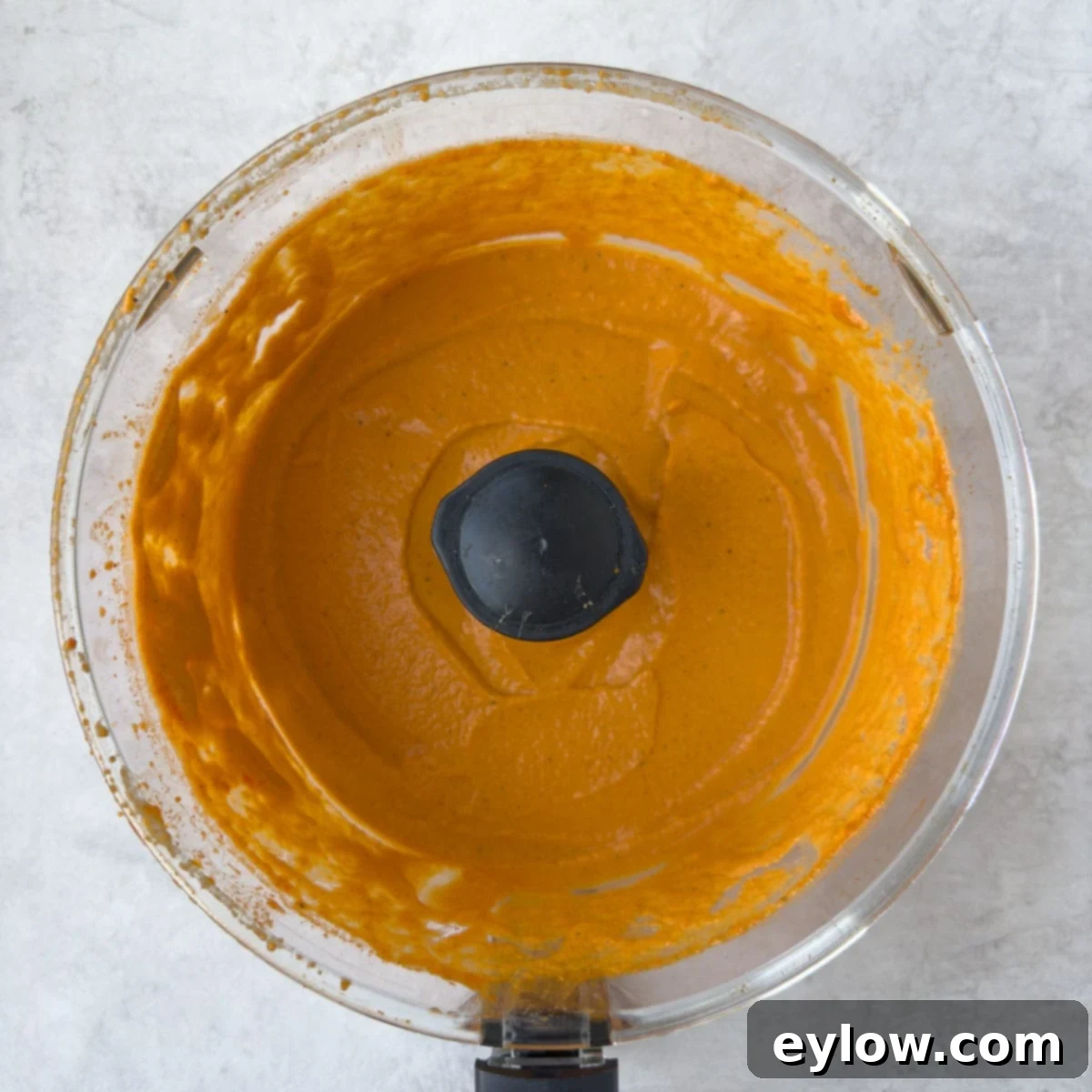 A top-down view of a food processor workbowl filled with perfectly creamy, bright orange roasted red pepper hummus, showcasing its smooth and inviting texture.