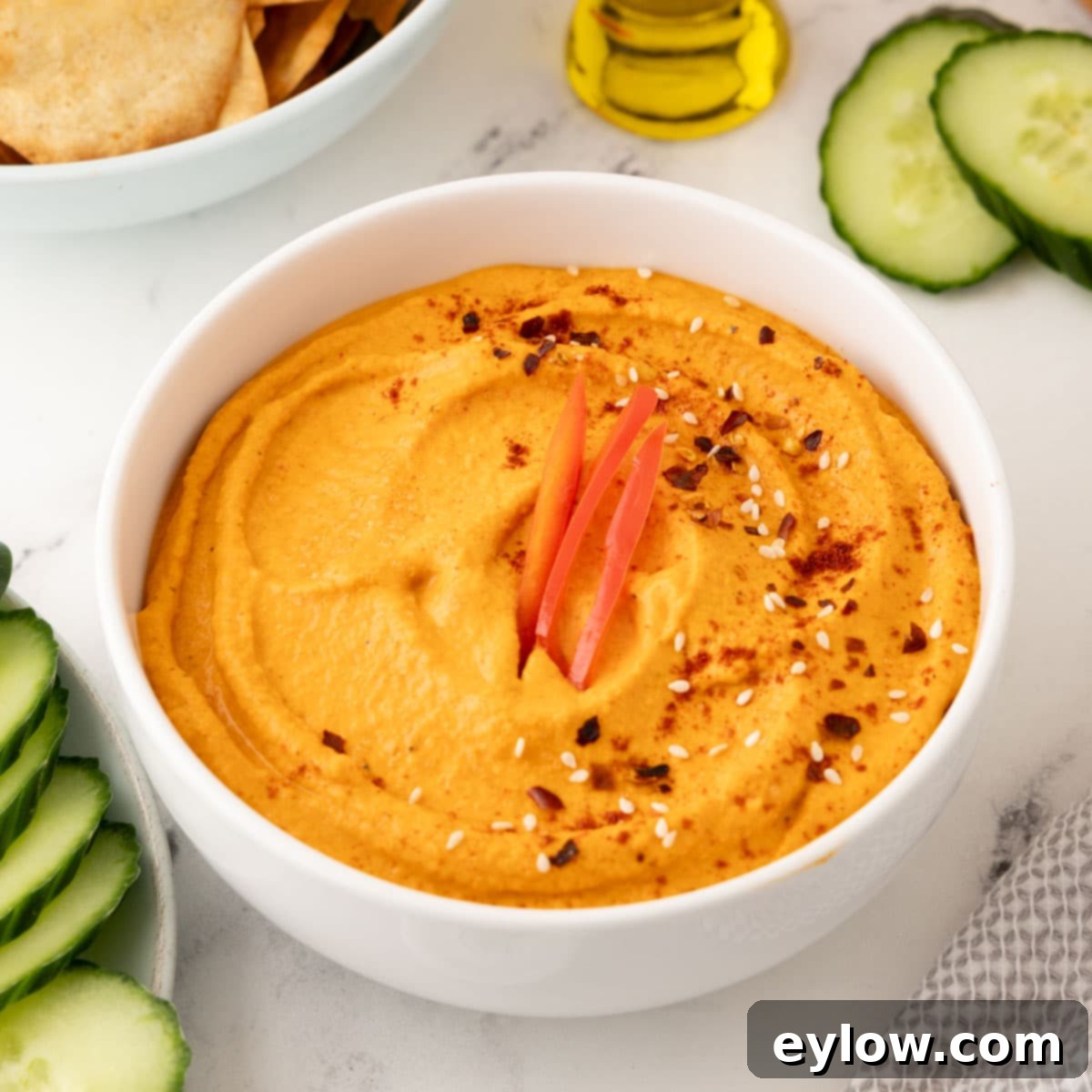A elegant white ceramic bowl filled with vibrant roasted red pepper hummus, beautifully drizzled with extra virgin olive oil and lightly sprinkled with fresh herbs, positioned as a inviting appetizer.