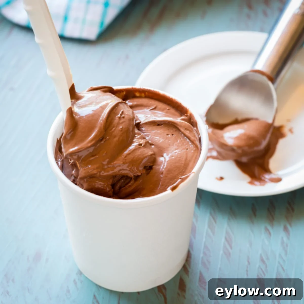 A white paper container of creamy dark chocolate ice cream on a blue counter with a scoop.