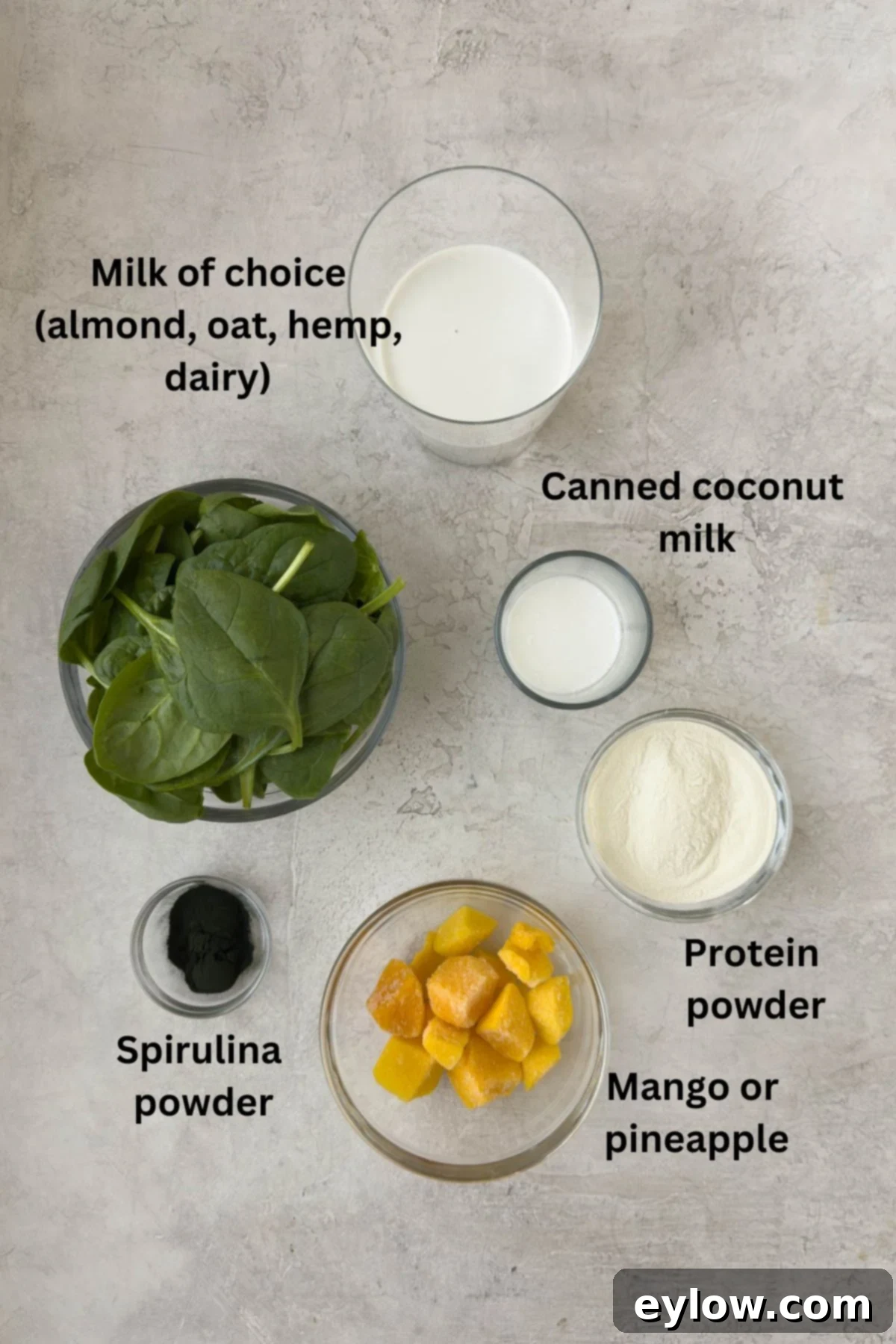Revitalizing Green Detox Smoothie 3 Various ingredients for a green detox smoothie, including almond milk, coconut milk, spinach, pineapple, and spirulina powder, arranged in bowls around a blender.