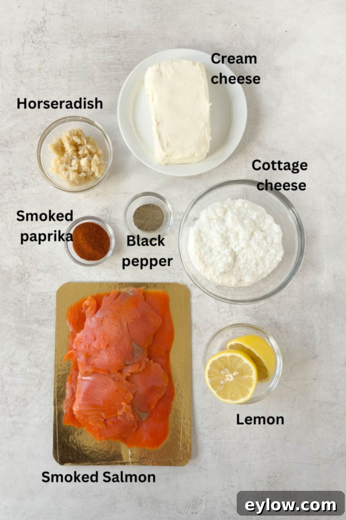 Ultimate Creamy Smoked Salmon Dip 3 Ingredients for smoked salmon dip laid out in glass prep bowls on a sleek gray counter, including salmon, cheeses, lemon, and spices.