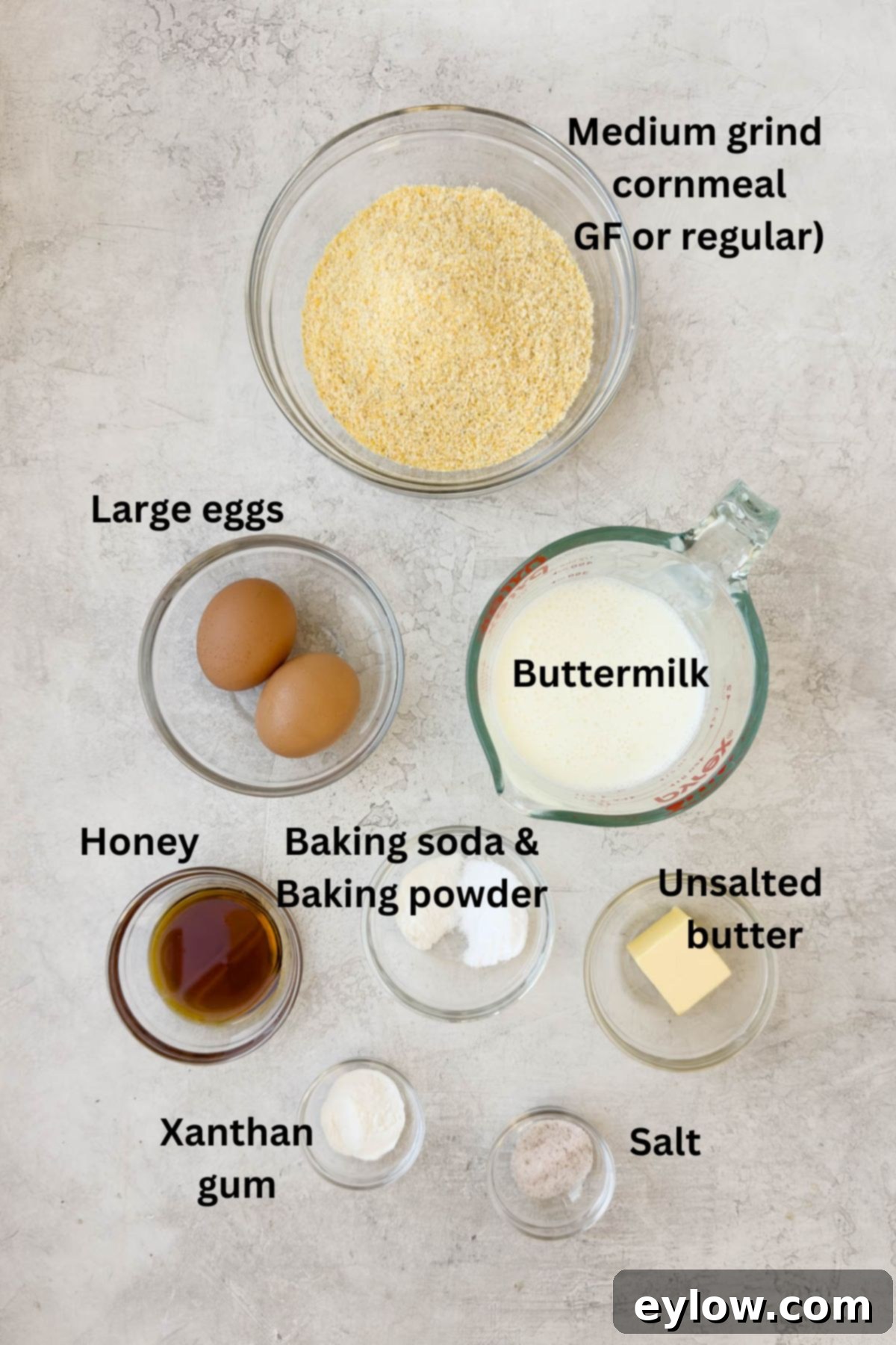 Ingredients in prep bowls on a kitchen counter for making cornbread, showing cornmeal, butter, honey, and other components.