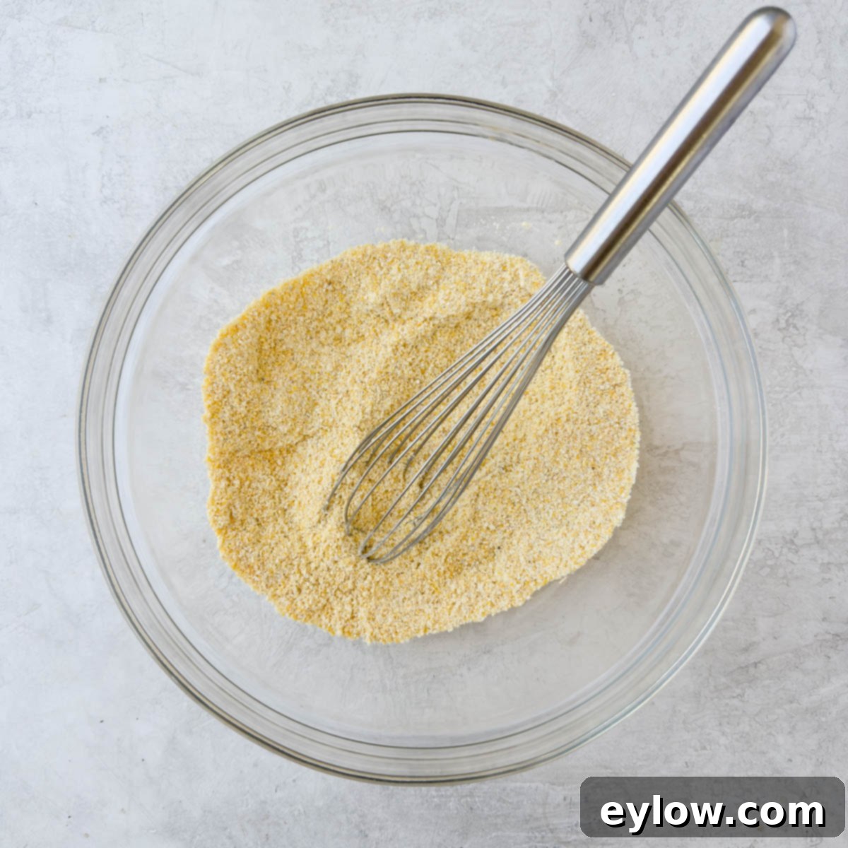 A glass mixing bowl with cornmeal and dry ingredients, ready to be whisked with a silver whisk.