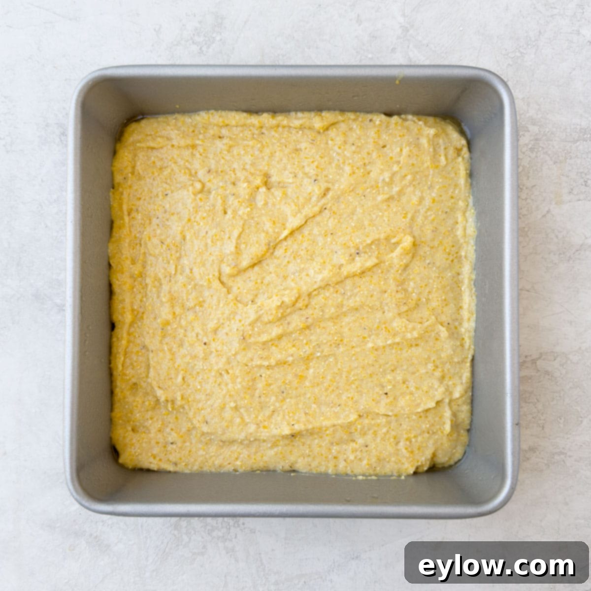 Smooth cornbread batter poured into a square silver baking pan, ready to be placed in the oven.