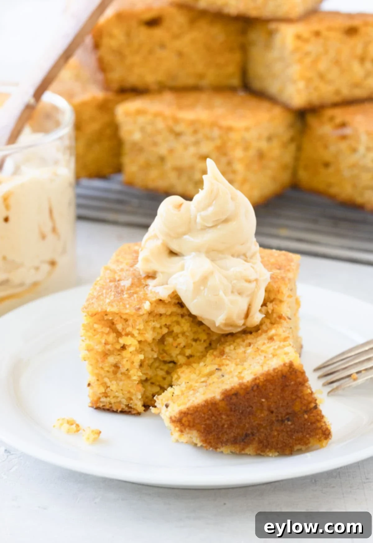 A piece of golden cornbread topped with a generous smear of homemade maple butter on a white plate with a fork.