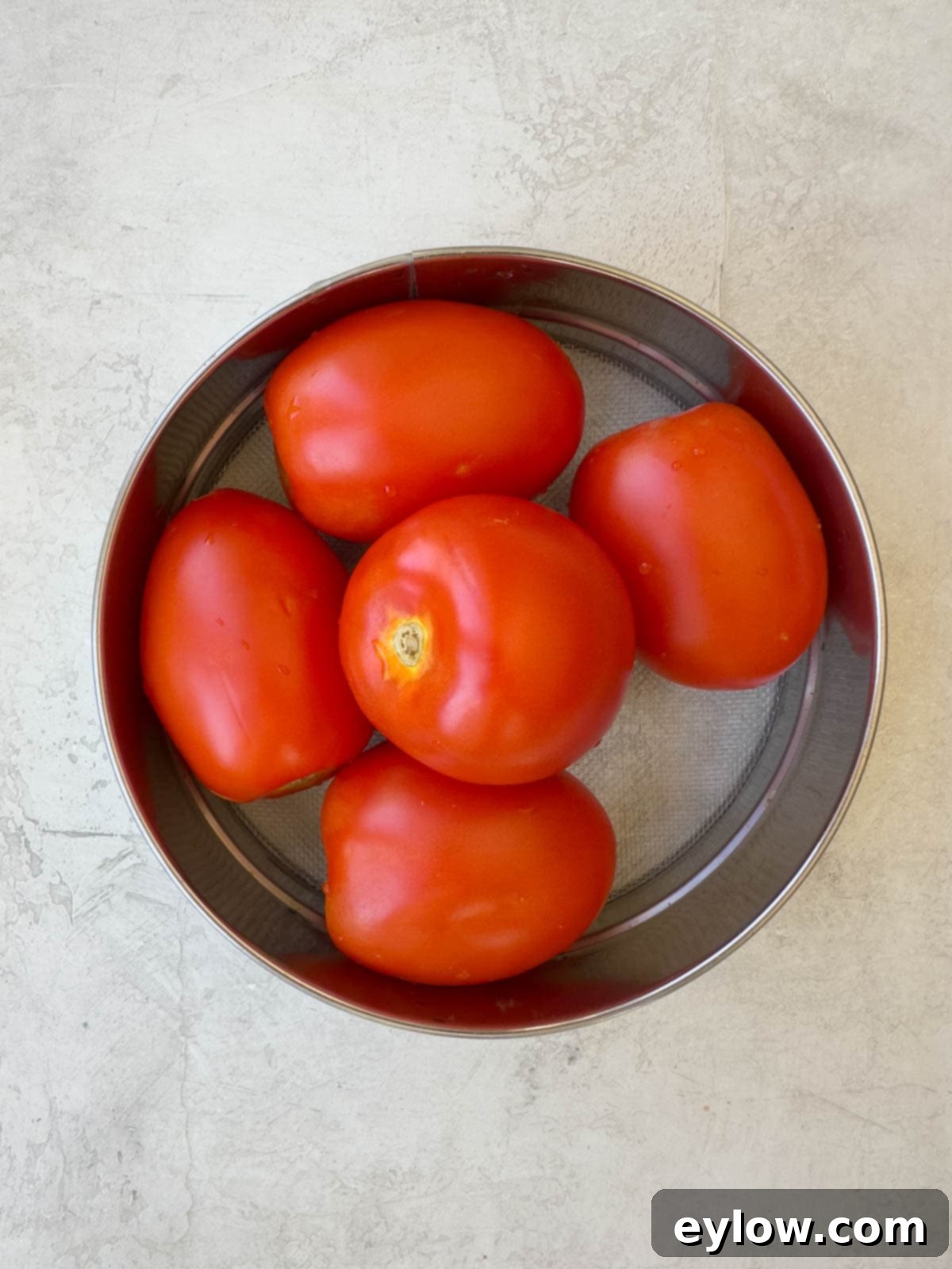 A collection of whole, ripe Roma tomatoes resting in a round colander after being freshly washed, ready for preparation.