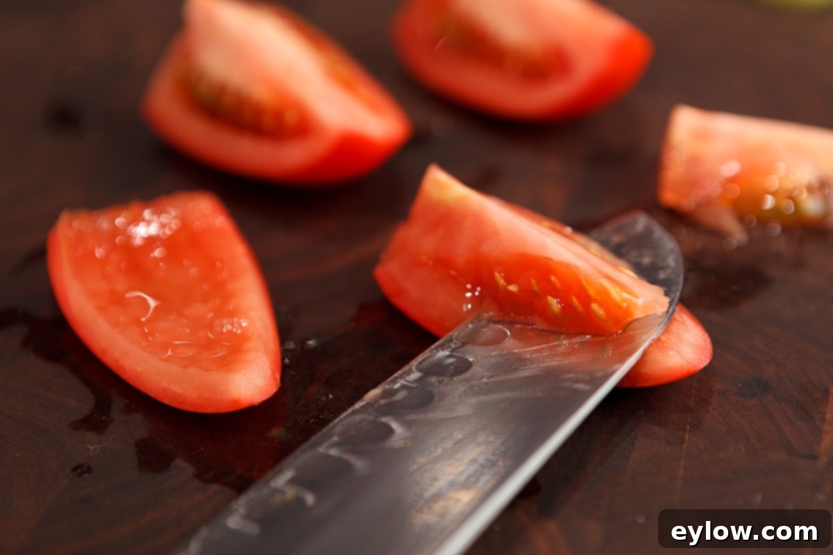 A sharp chef's knife expertly removing the seedy, pulpy centers from quartered Roma tomatoes on a walnut cutting board.