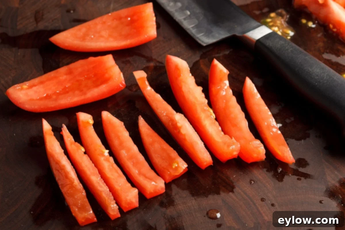Cleaned Roma tomato quarters are cut into uniform long strips on a dark brown cutting board, poised for crosswise dicing.
