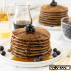 Stacked buckwheat pancakes on a white plate with syrup and blueberries.