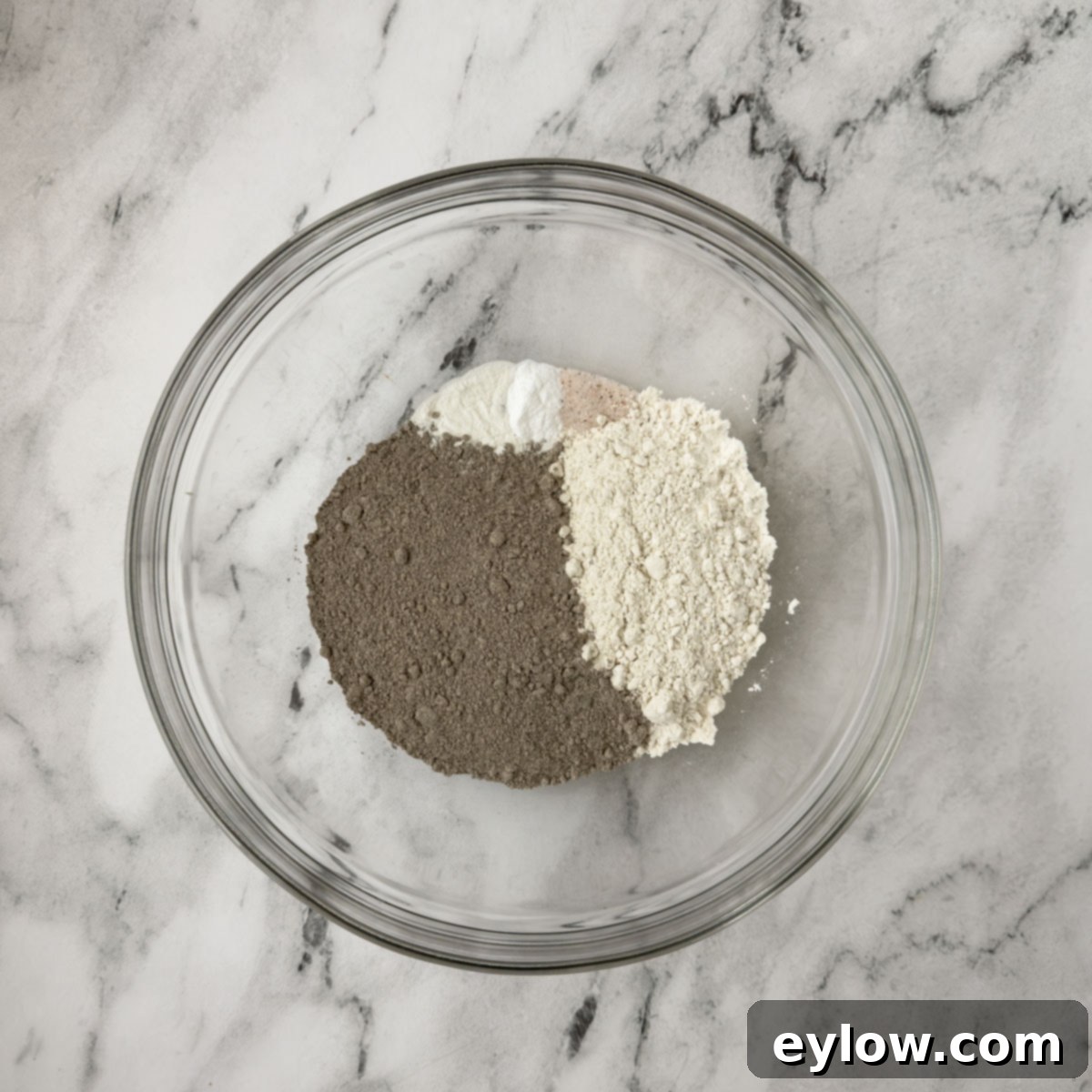 Dry ingredients for pancakes, including buckwheat and regular flour, baking powder, baking soda, and salt, perfectly combined in a clear glass mixing bowl.